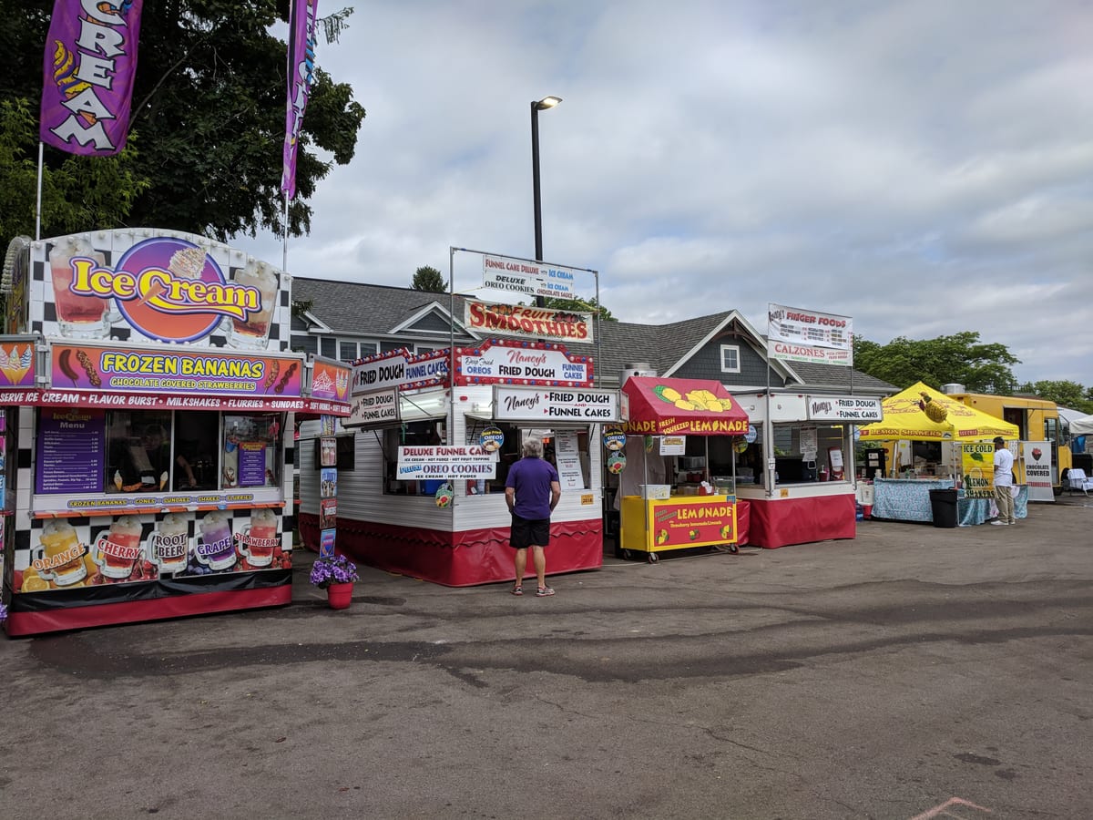 Row of brightly painted festival food stands selling ice cream, frozen bananas, fried dough, funnel cakes and lemonade under a cloudy sky