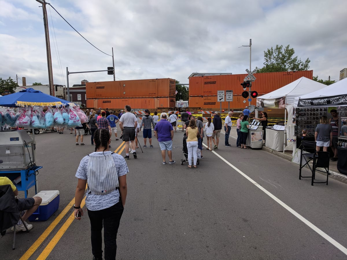 Festival crowd on North Main Street waiting at a lowered railroad crossing while a CSX freight train of orange stacked containers passes through
