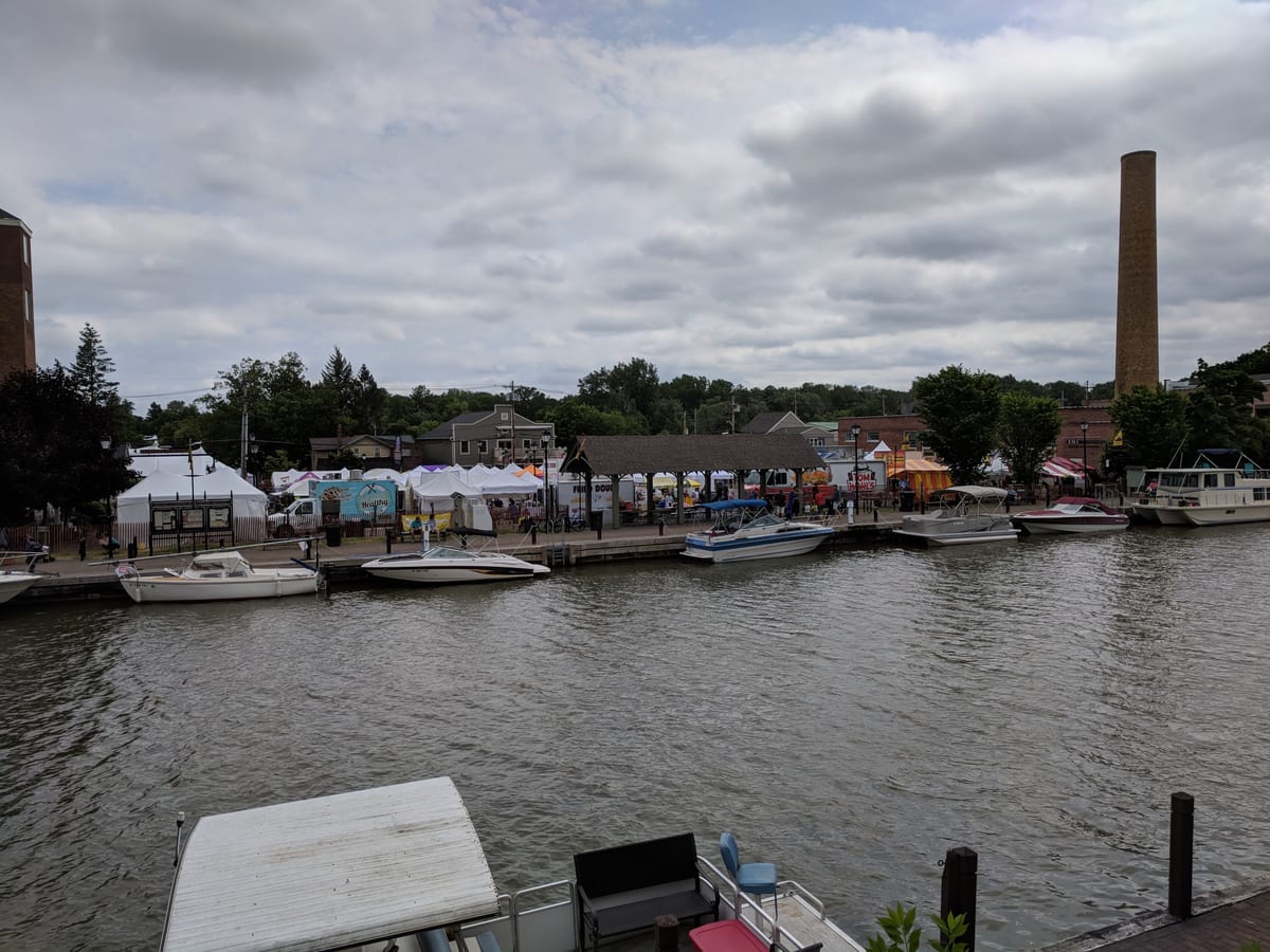 White festival tents and a wooden pavilion along the north bank of the Erie Canal, with motorboats tied up at the marina and a tall smokestack behind