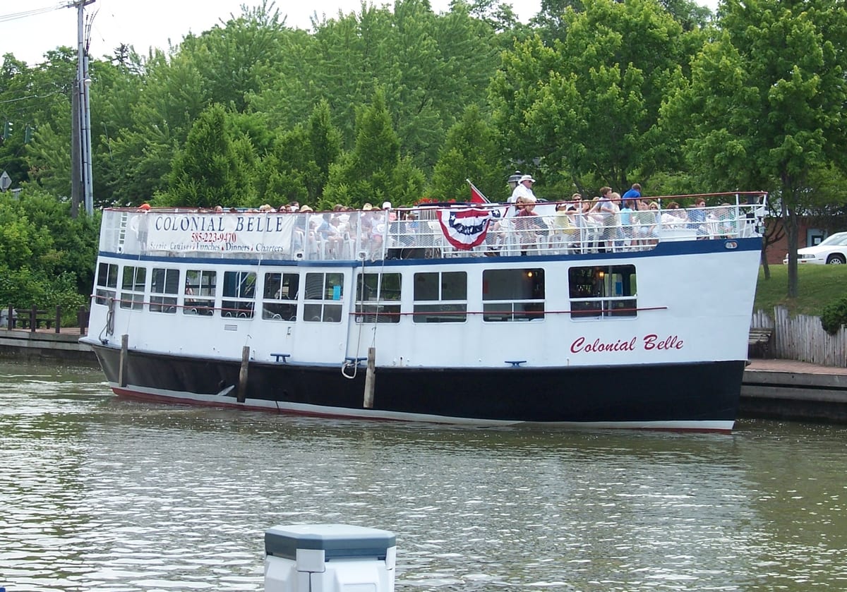 White tour boat with a dark hull and Colonial Belle lettered on its side, passengers on the open upper deck, moving along a tree-lined canal wall
