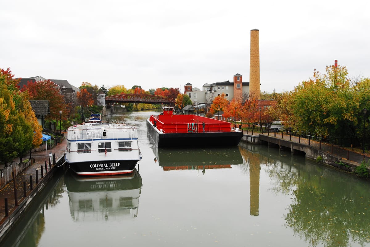 Autumn view down the Erie Canal with the Colonial Belle docked on the left, a red barge in mid-channel, a tall brick smokestack, and the lift bridge in the distance