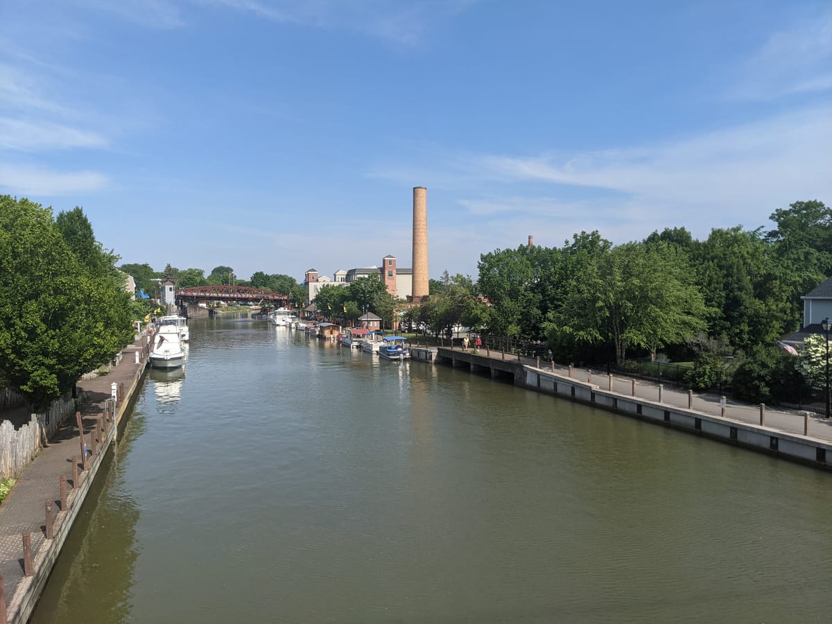 Wide, calm Erie Canal under a blue summer sky, lined by brick walkways and moored boats, leading west to the red lift bridge and a tall brick smokestack