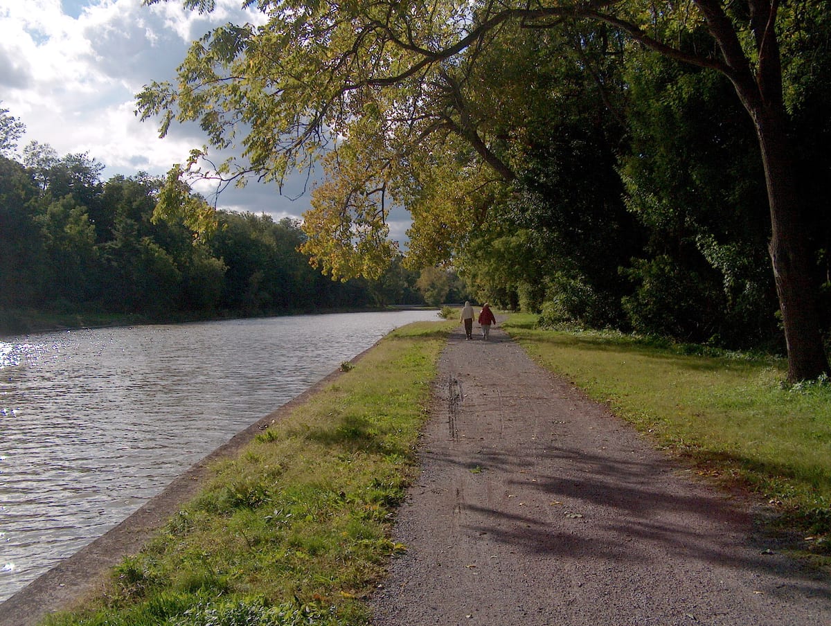 Two walkers on a gravel towpath beside the Erie Canal under arching trees, the water on the left reflecting a bright sky