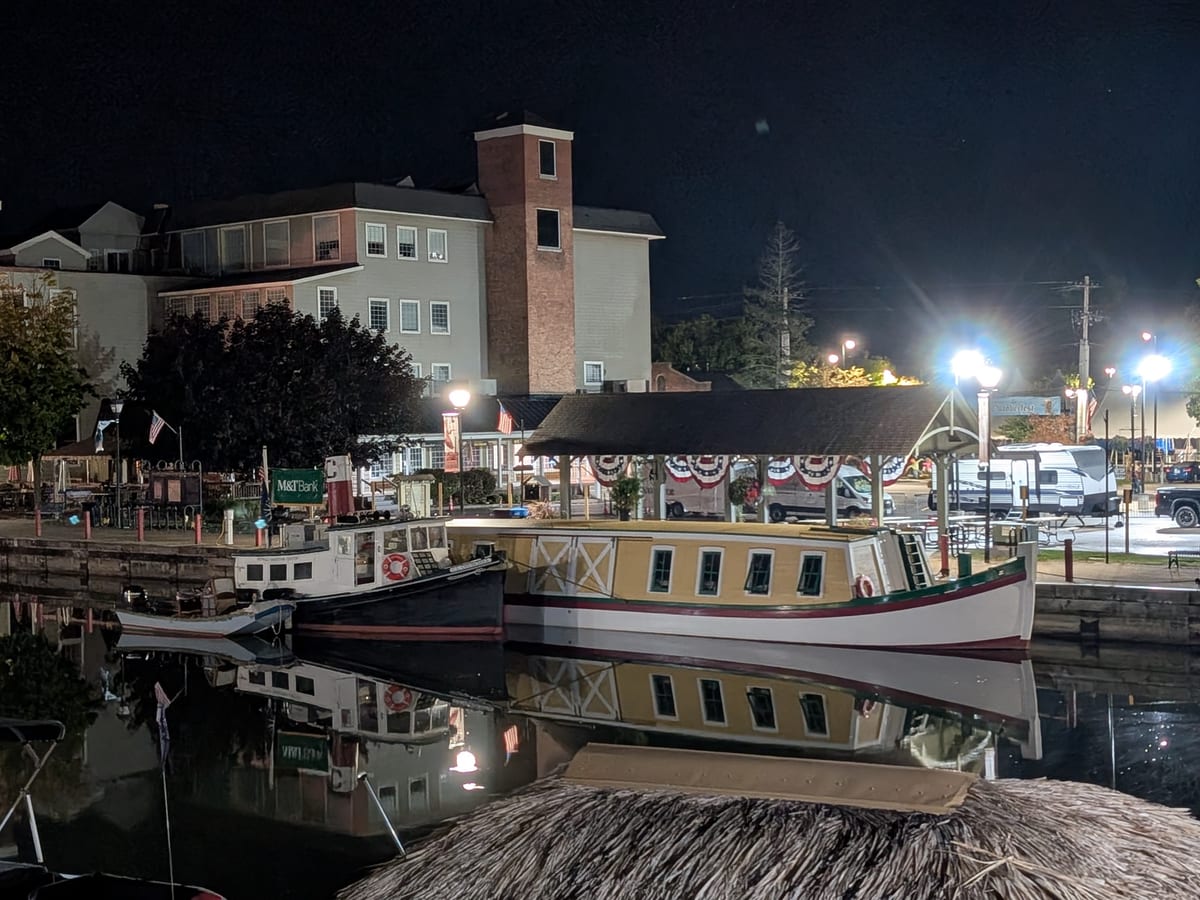 Fairport canal docks at night with a replica packet boat and a small tug reflected in black water, lit lamp posts and a pavilion behind