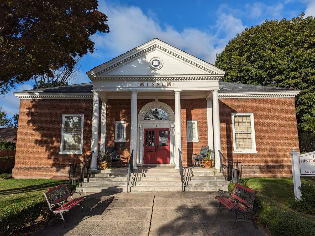 Red-brick building with a white columned portico, red double doors and the word MUSEUM above the entrance, with iron benches on the front walk