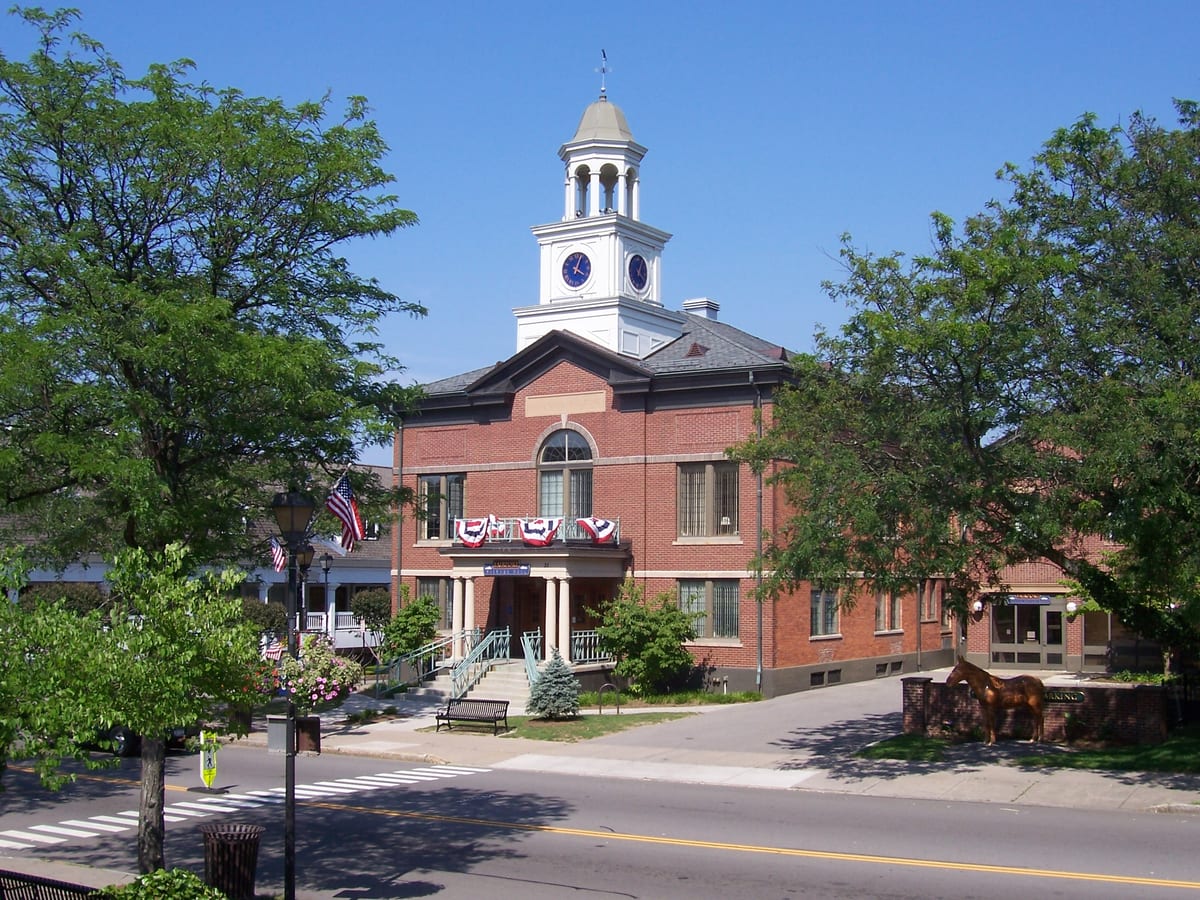 Red-brick village hall with a white clock cupola, red, white and blue bunting over the entrance portico and a bronze horse sculpture beside the side walkway