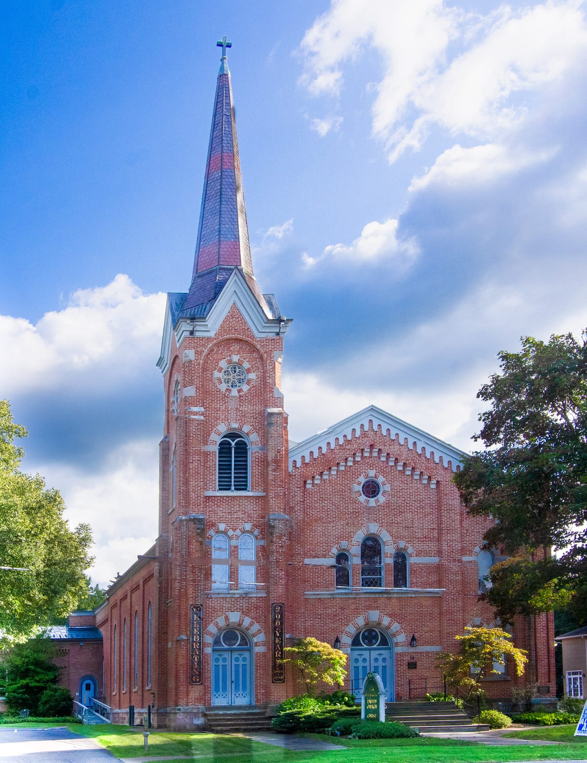 Red-brick church with a tall slate spire, round rose windows and blue arched doors behind a green lawn under a bright partly cloudy sky