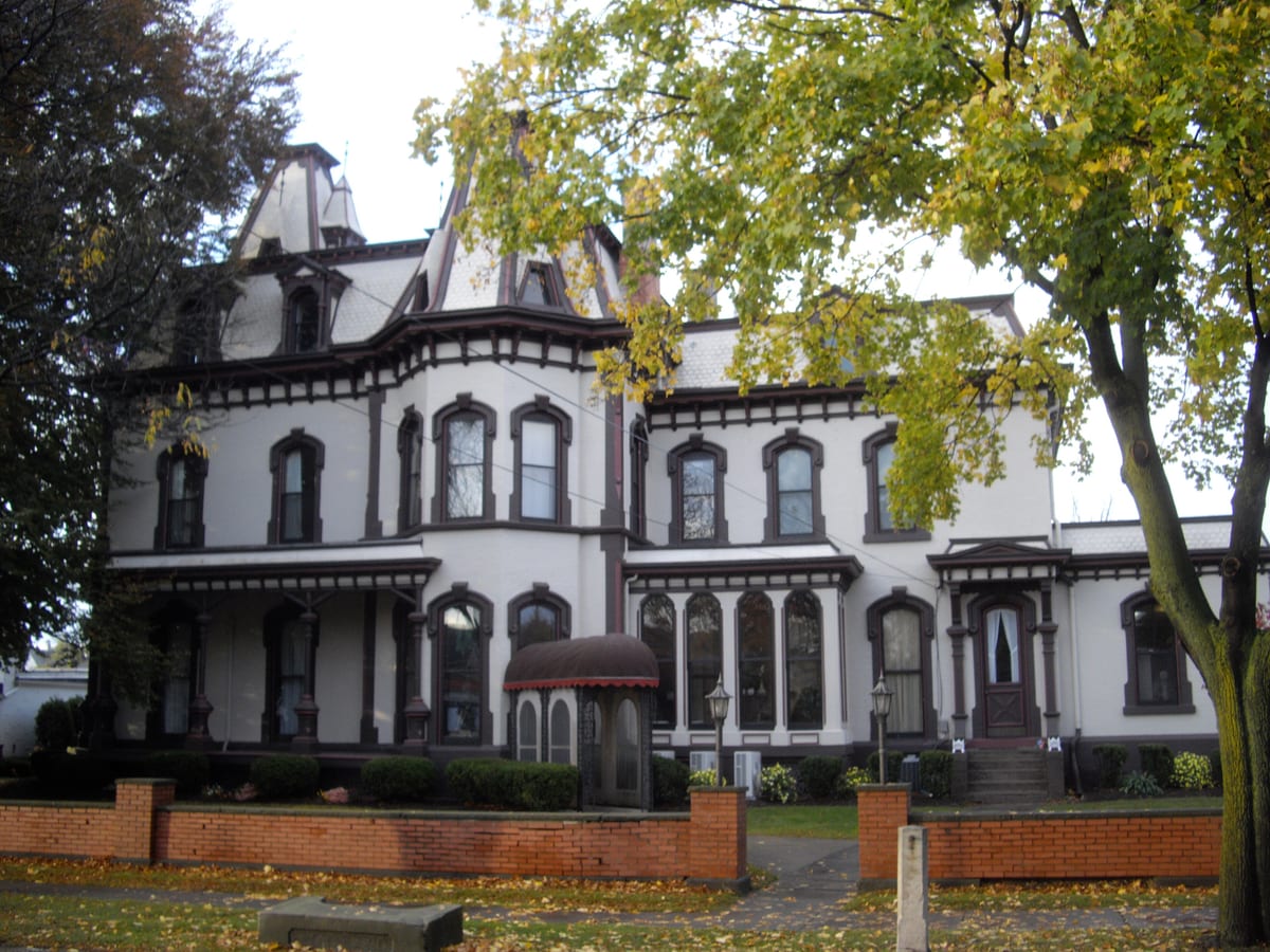 Large white Second Empire house with a mansard roof, arched dark-trimmed windows and a covered side entrance behind a low red-brick wall, autumn leaves on the lawn