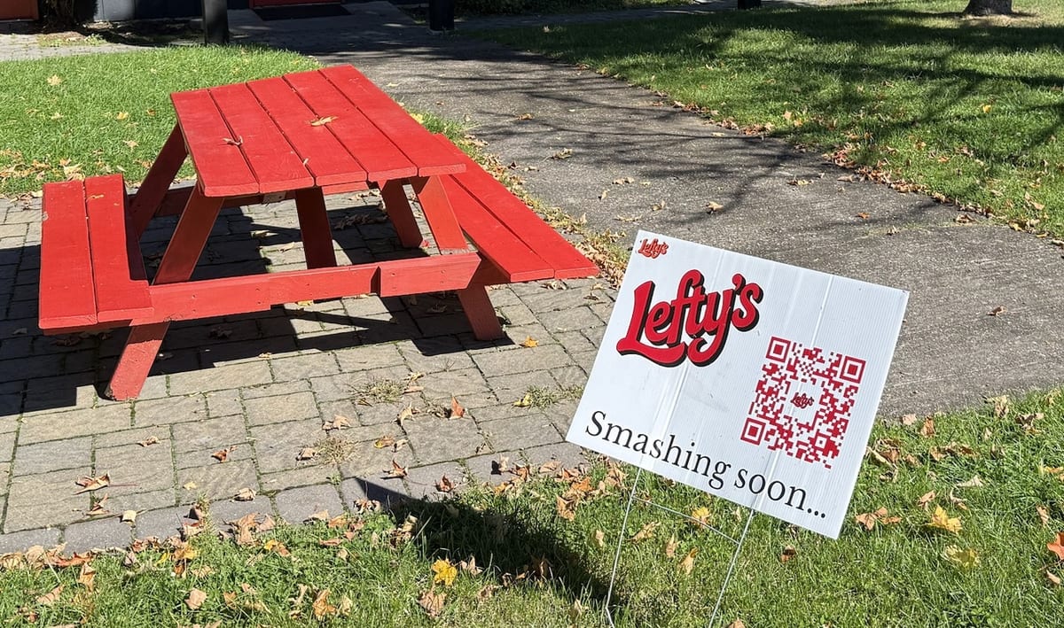 A white yard sign with a red Lefty's logo, a QR code and the words Smashing soon, beside a red picnic table on a brick patio
