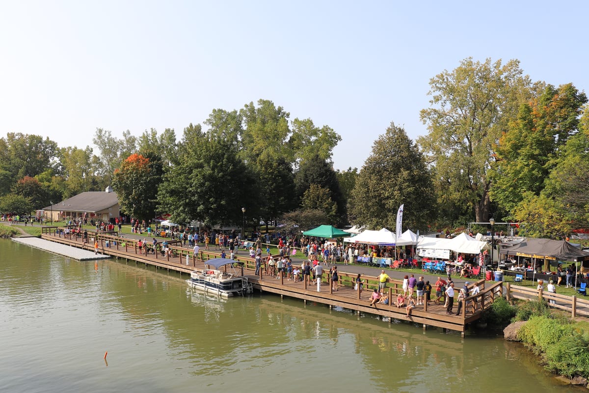 Crowds and white tents along a wooden dock beside the Erie Canal at the Lift Bridge Regatta, with a pontoon boat tied up and trees starting to turn