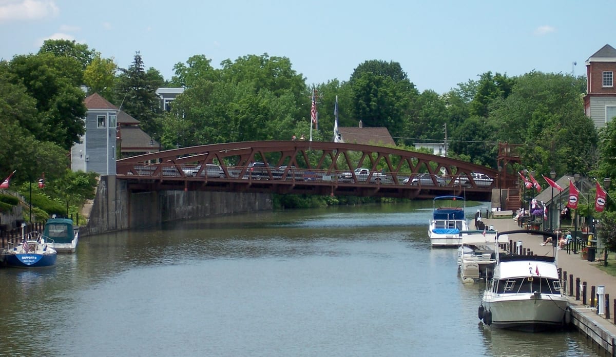 Red arched steel truss bridge with cars on its deck crossing the Erie Canal, pleasure boats tied up along a promenade hung with red pennants on the right