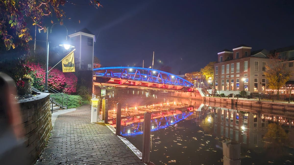 Lift bridge outlined in blue and red lights at night, reflected in the dark canal beside a lamp-lit brick walkway and a brick building with lit windows
