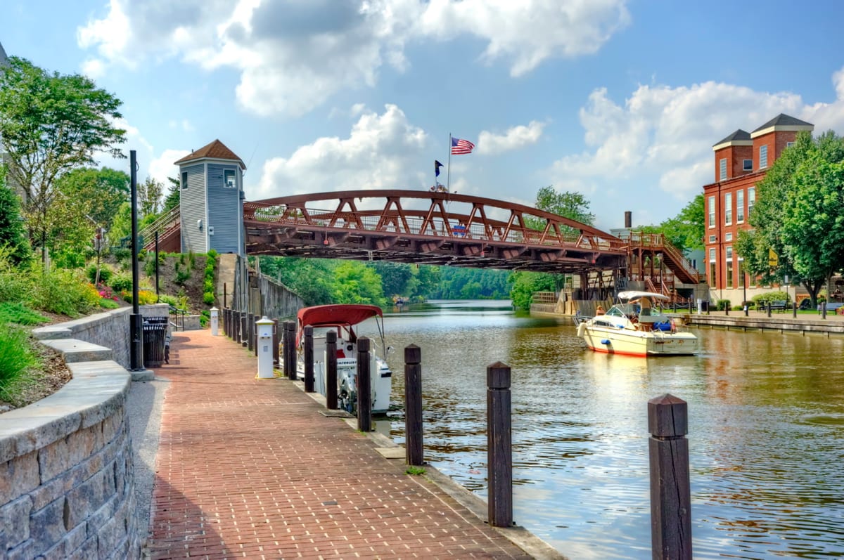 Rust-red steel truss lift bridge spanning the Erie Canal, seen from a brick walkway lined with wooden bollards, with moored boats and brick buildings along the far bank