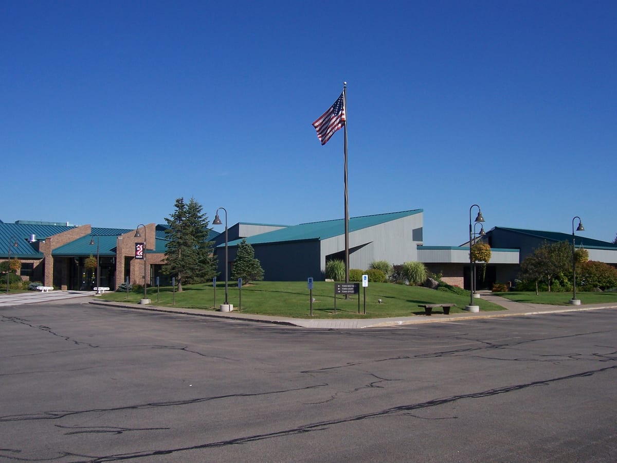 Low brick and gray-paneled municipal building with teal metal roofs and an American flag on a tall pole over the front lawn, a parking lot in the foreground