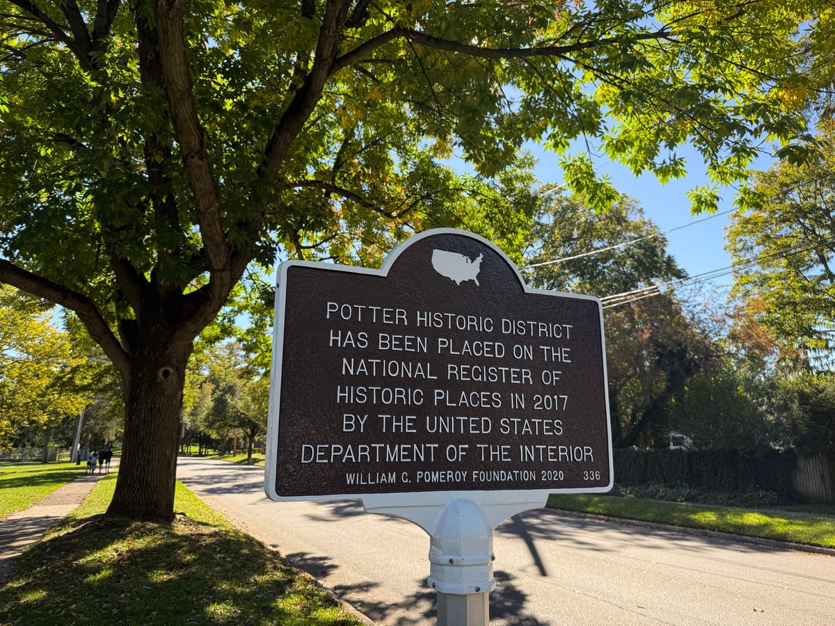A brown and white National Register marker for the Potter Historic District on a white post beside a quiet tree-lined street, with a large shade tree and a sidewalk on the left