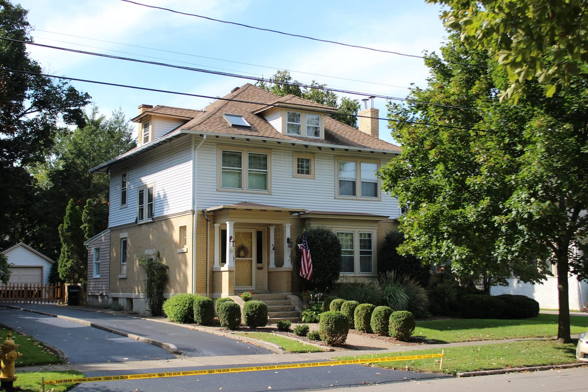 Two-story house with a cream brick ground floor, light siding above, a small columned porch and rounded clipped shrubs along a curving driveway