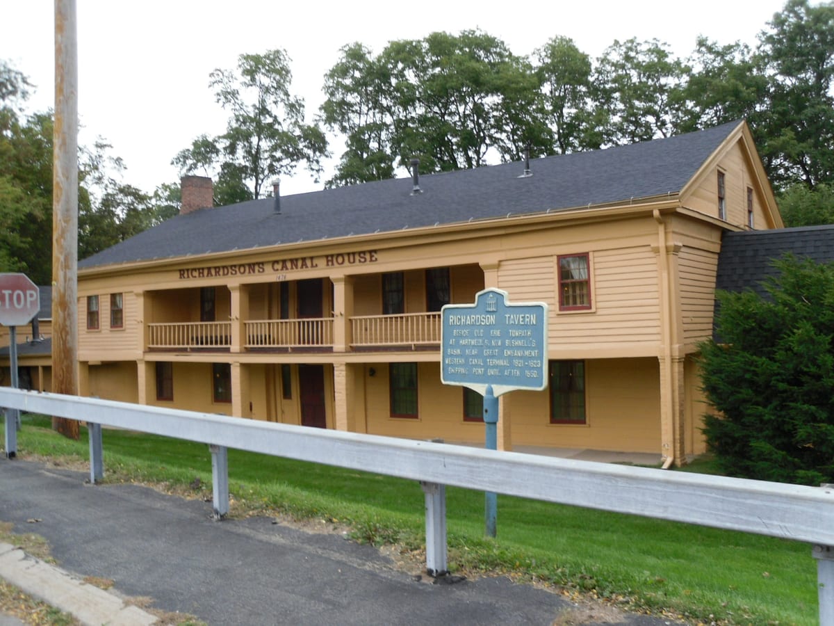 Long two-story yellow clapboard tavern with a full-length balcony and a blue historic marker on the lawn beside a road guardrail