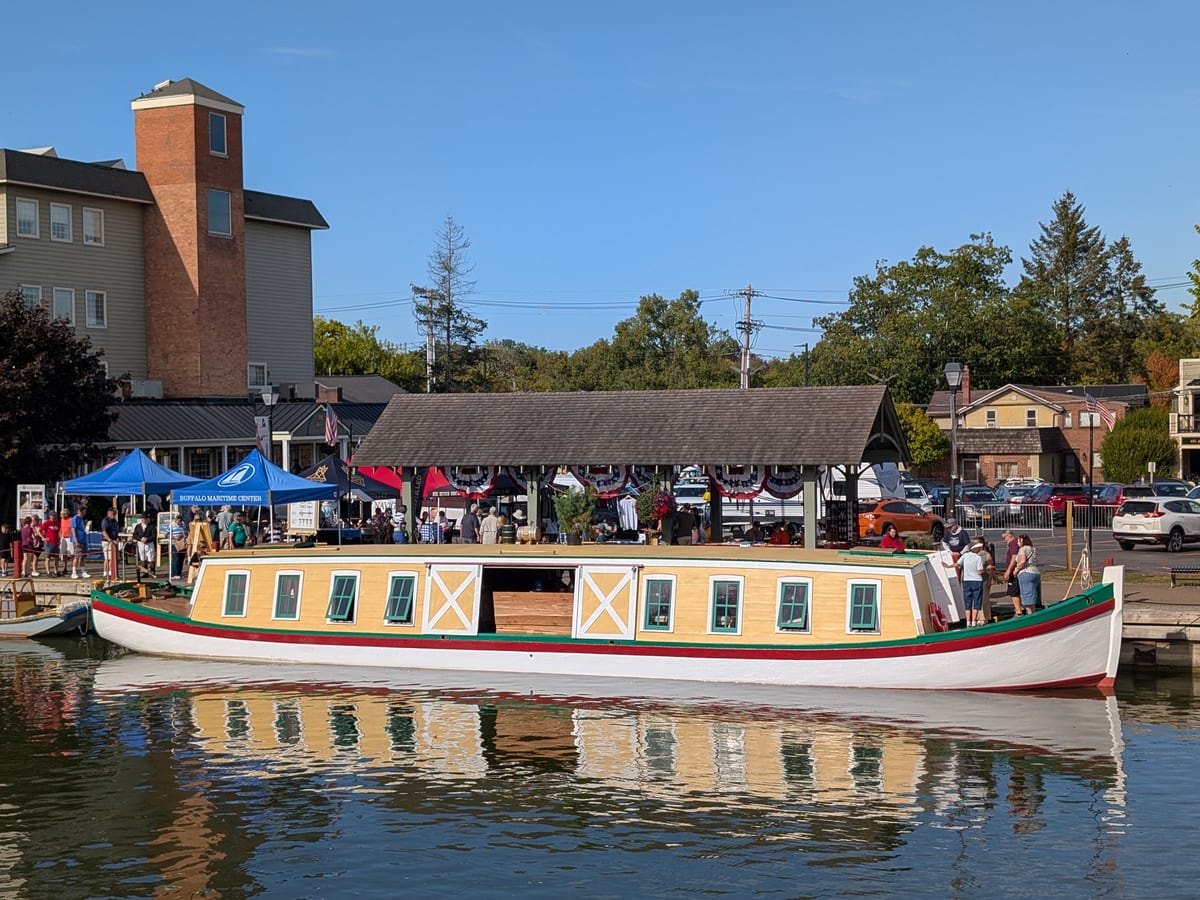 Cream and green replica canal packet boat tied up at the Fairport docks beside a shingled pavilion hung with red, white and blue bunting, visitors on the dock