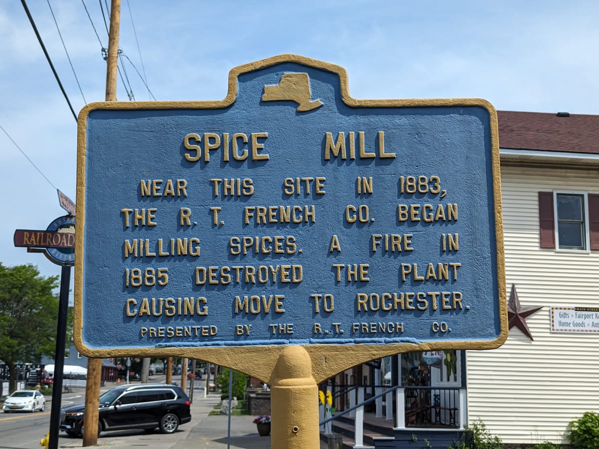 Blue and gold roadside marker reading Spice Mill on North Main Street, with a Railroad Street sign and shop fronts behind it