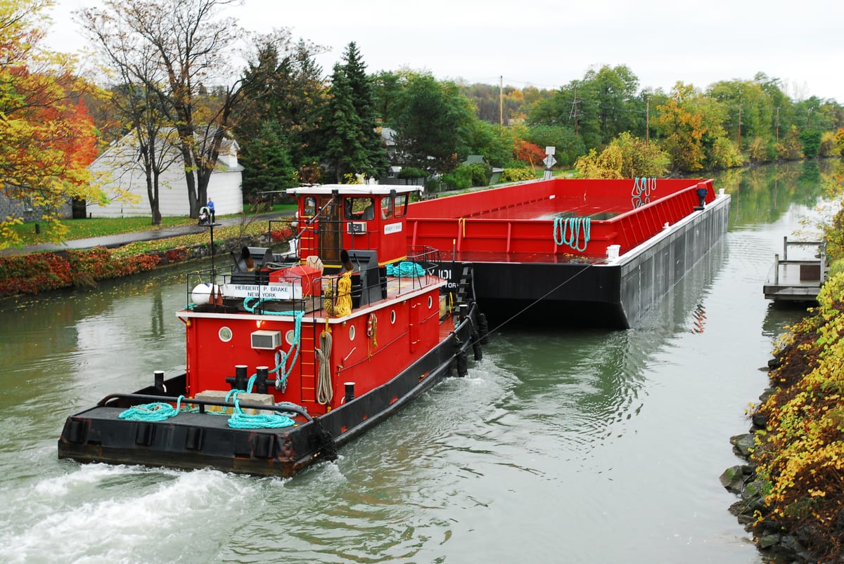 Bright red tugboat pushing an empty red barge east along the Erie Canal past orange and yellow autumn trees and a white house on the bank